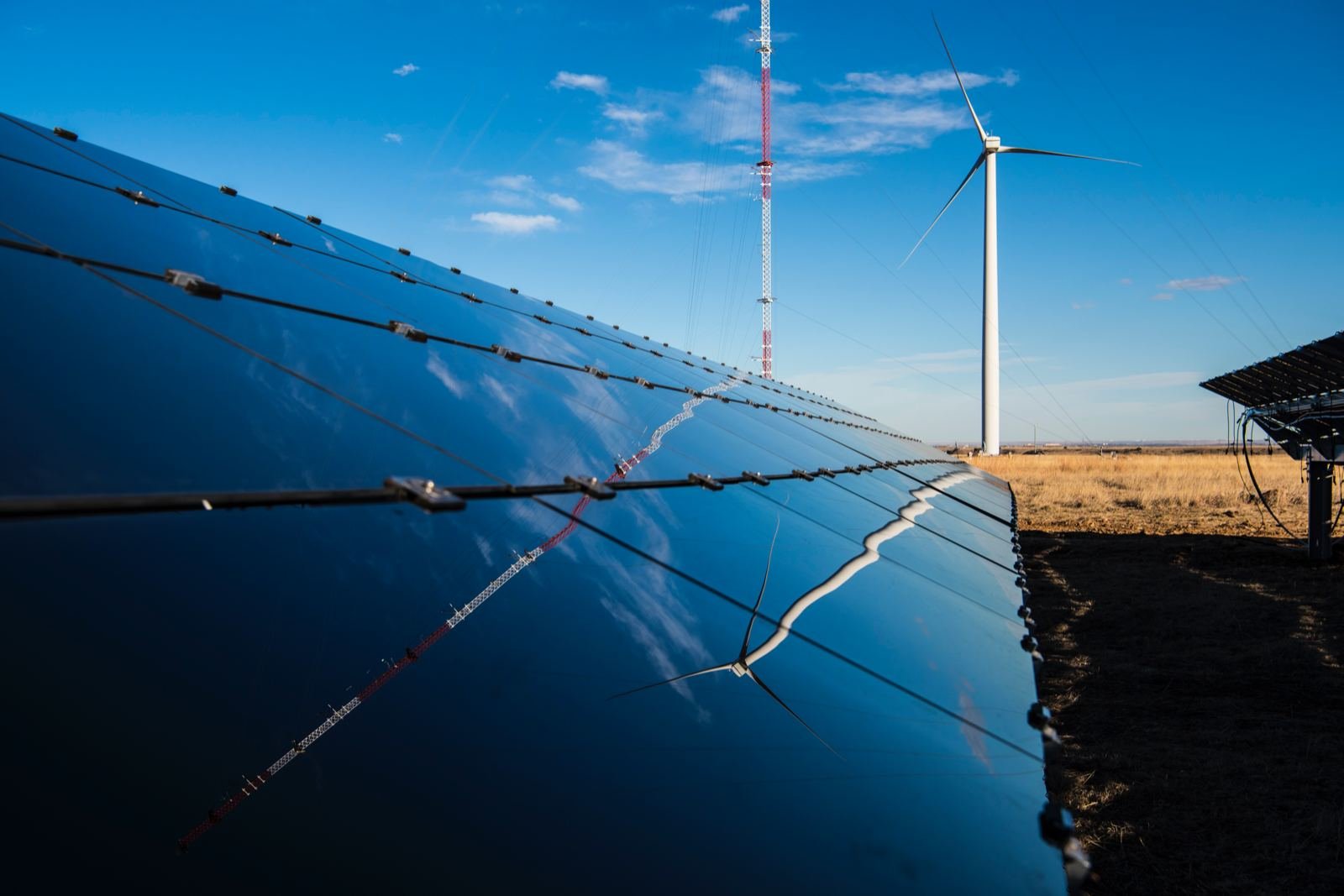 Row of solar panels on open grassland, reflecting a wind turbine and a tall measurement mast that stand behind them under a blue sky